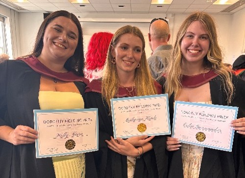 three ladies in graduation gowns holding prize certificates